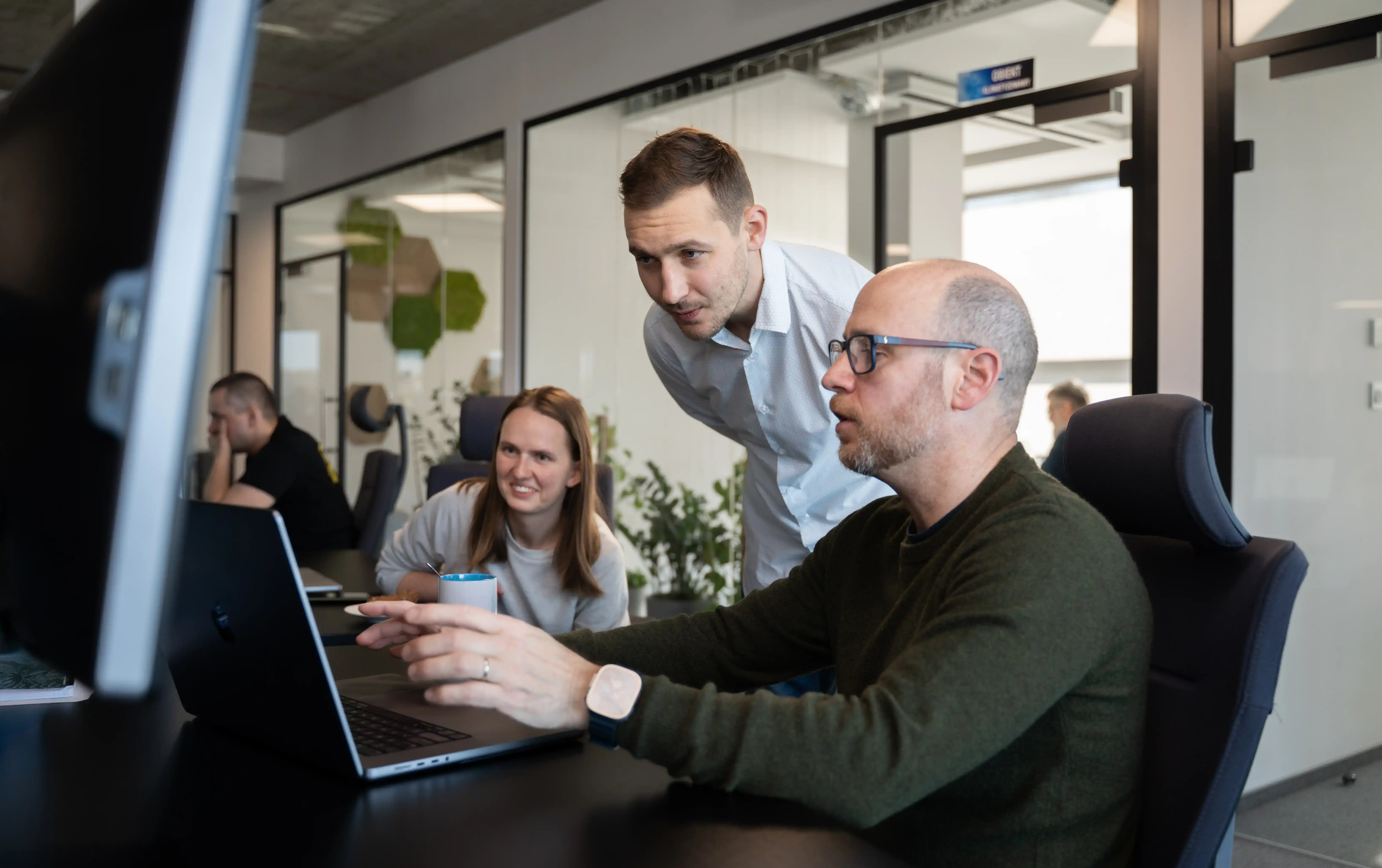 Three people in an office sit and stand around a laptop.